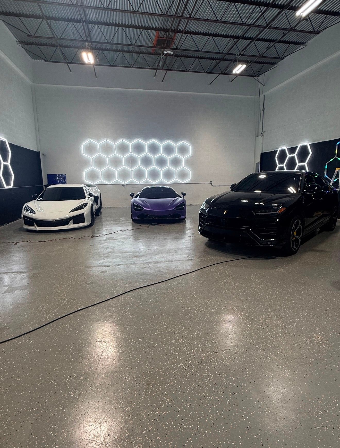 A white Corvette, a purple McLaren and a black Lamborghini Urus parked under hexagon lights in the fleet garage